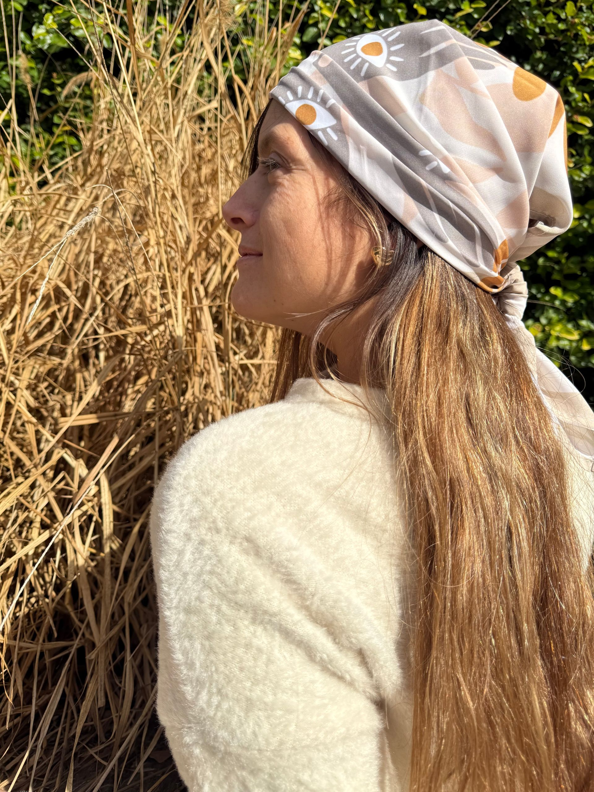 Mujer con cabello largo y castaño que lleva un pañuelo estampado en la cabeza, parada al aire libre cerca de pasto alto.