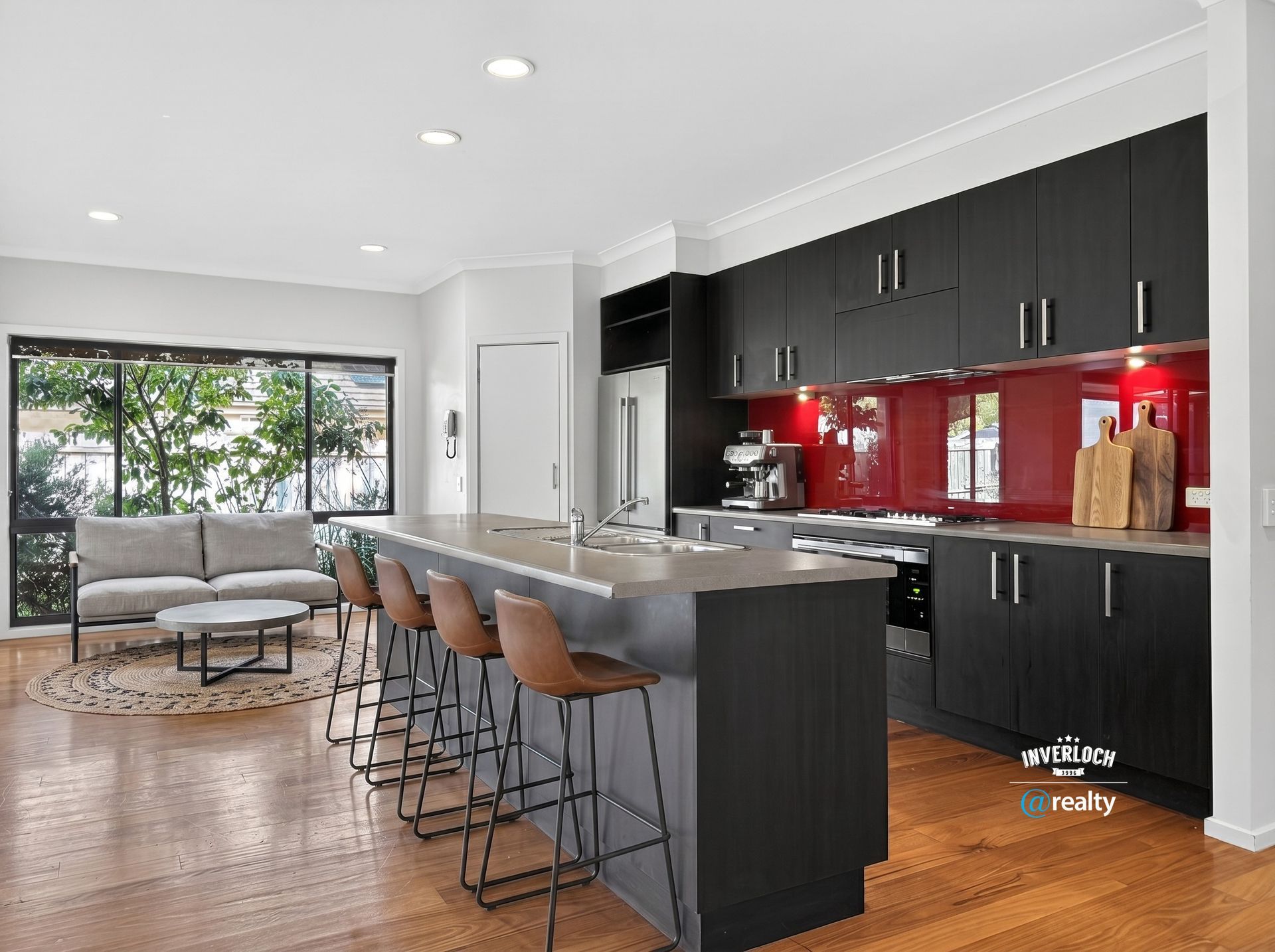 Modern open-plan kitchen with dark cabinets, red backsplash, breakfast bar, stools, and a gray sofa by large windows