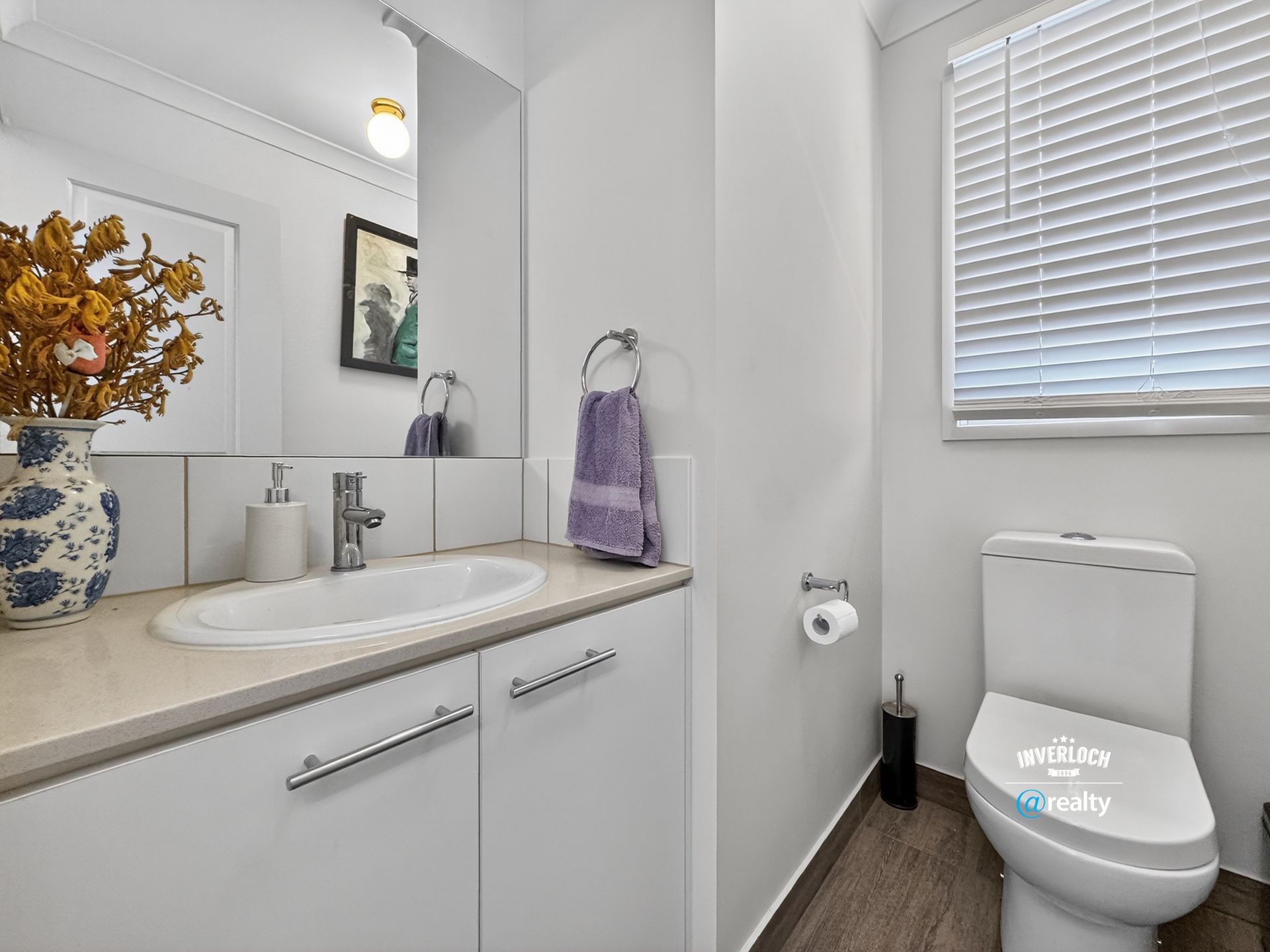 White bathroom with a sink, toilet, and window with blinds; flowers in a vase.