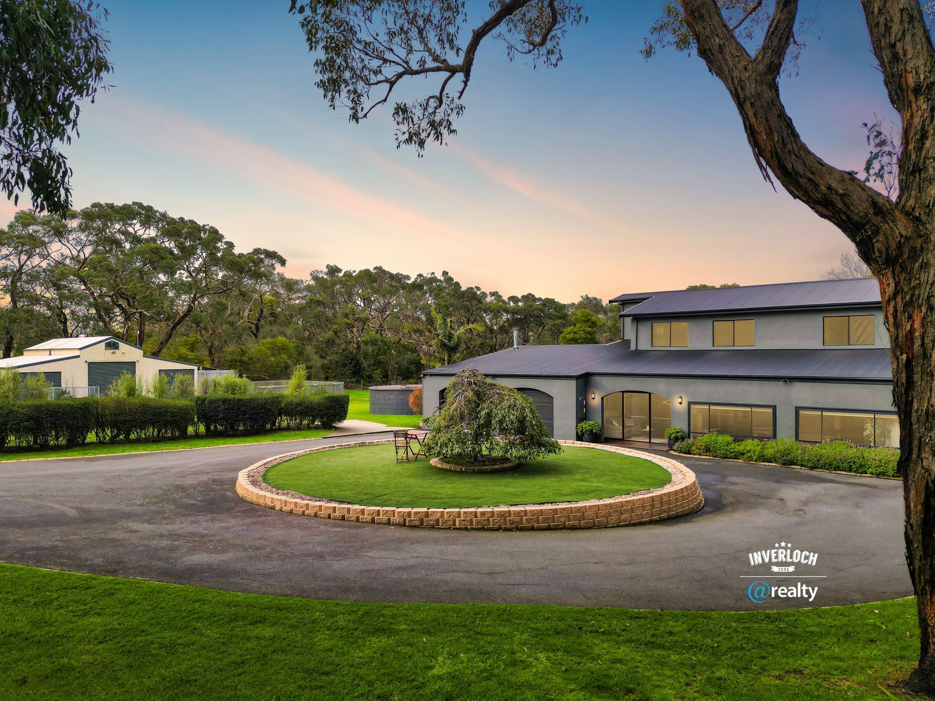 Large gray house with circular driveway and manicured lawn, trees.