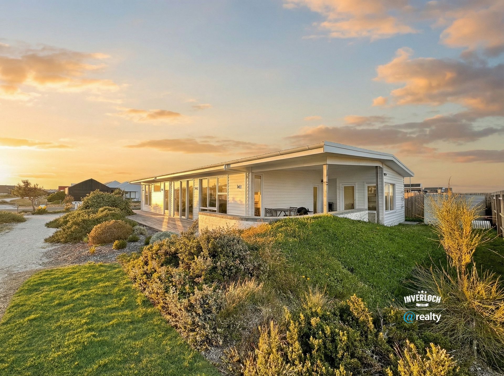 A white, modern, single-story beach house sits amidst coastal dunes and greenery at sunset.