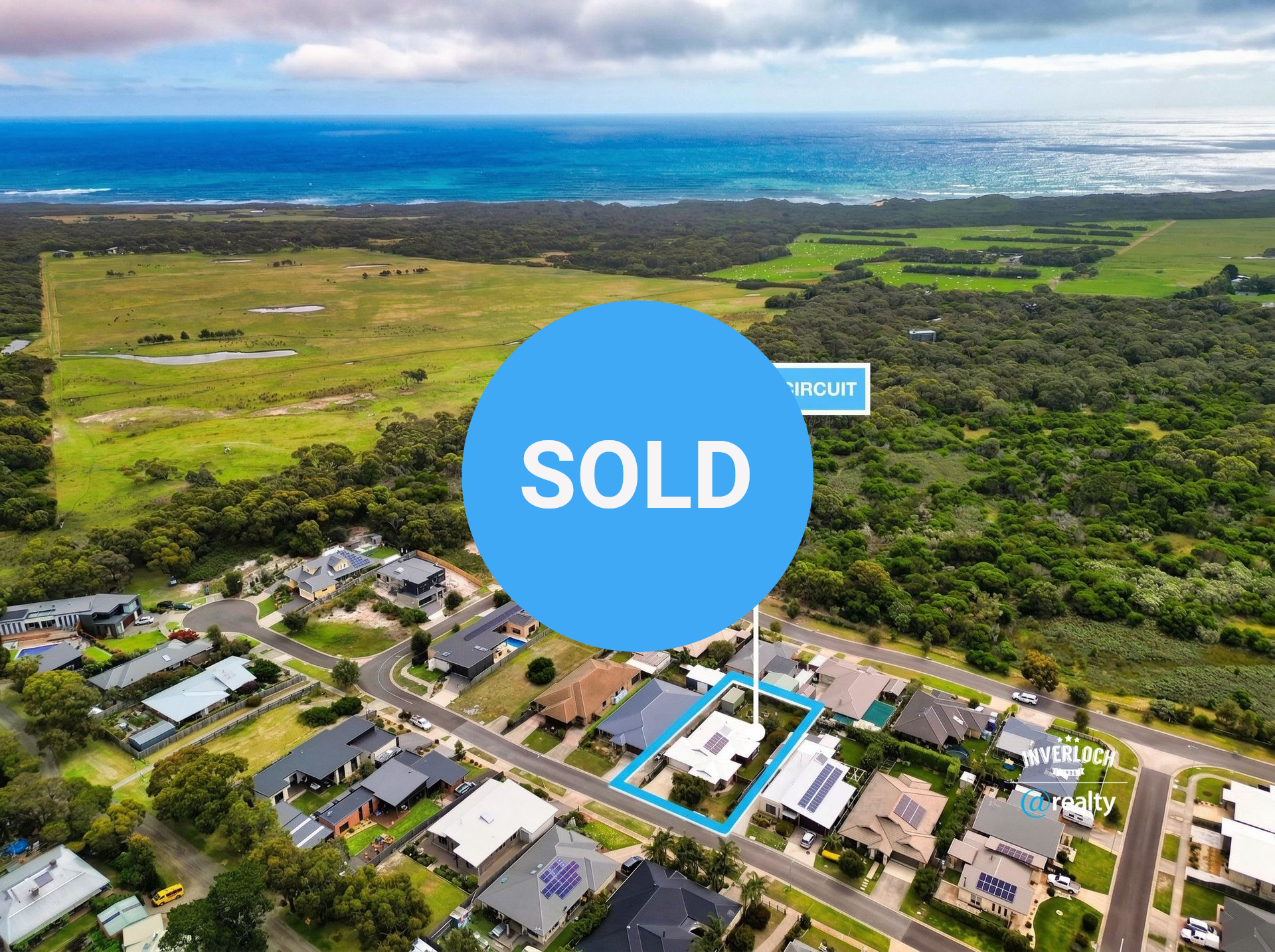 Aerial view of a suburban home highlighted in blue, surrounded by green fields and forest near the coast.