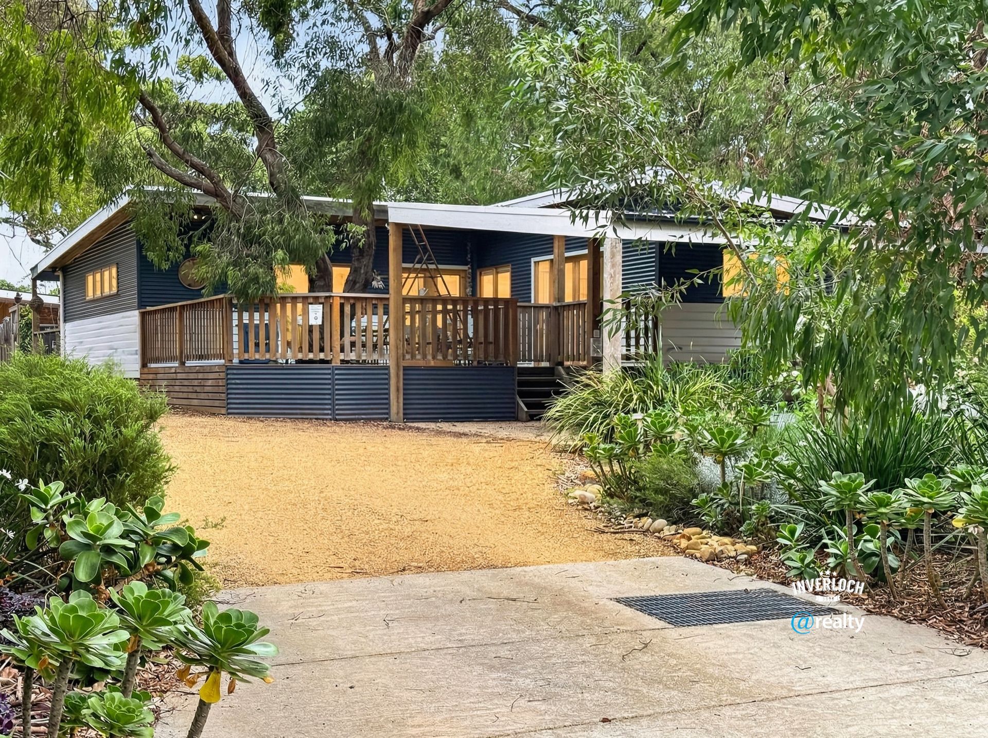 Modern black cabin with wooden porch, surrounded by trees and shrubs on a gravel driveway.