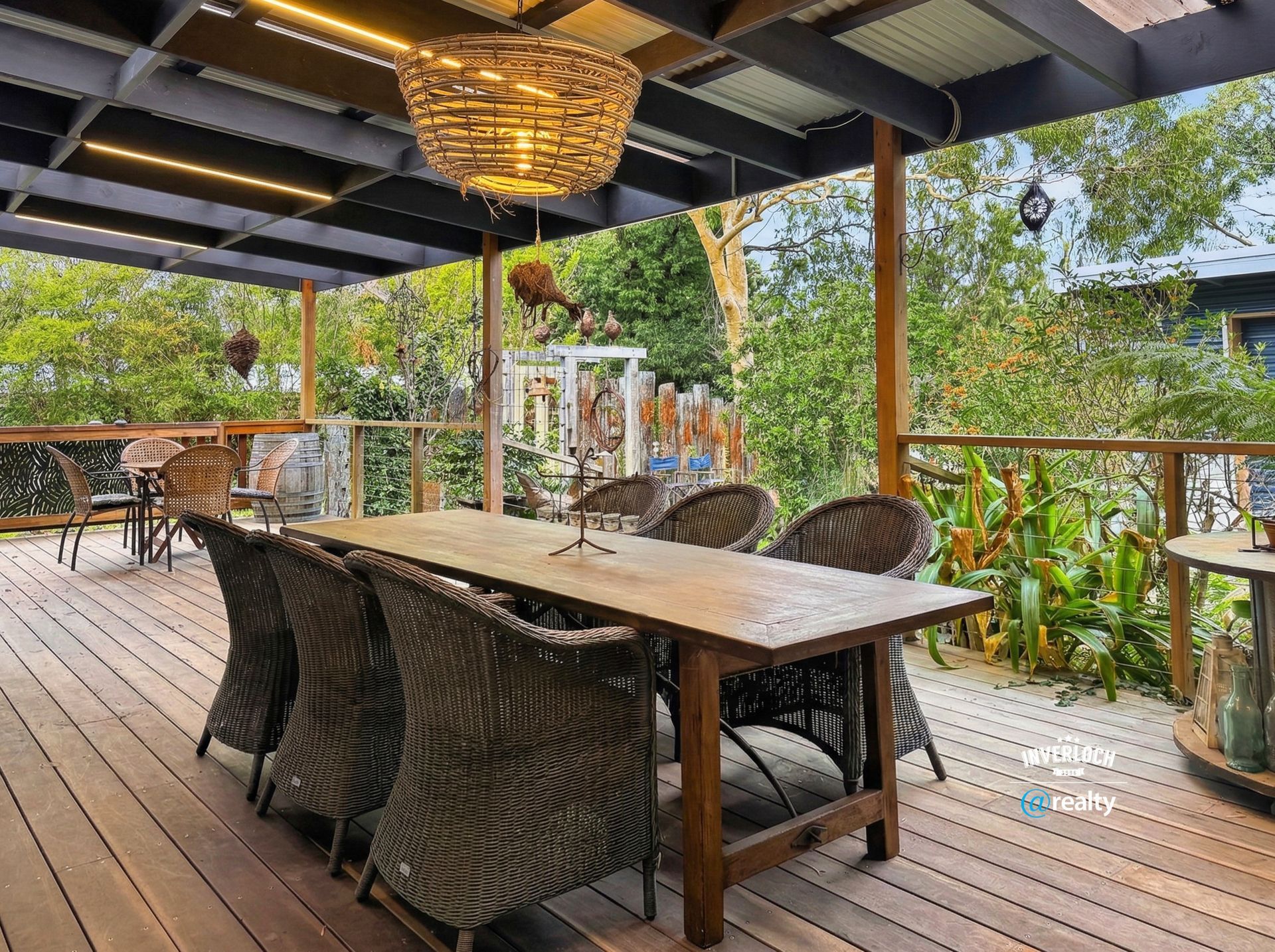 Covered wooden deck dining area with wicker chairs, long table, and lush greenery beyond the railing