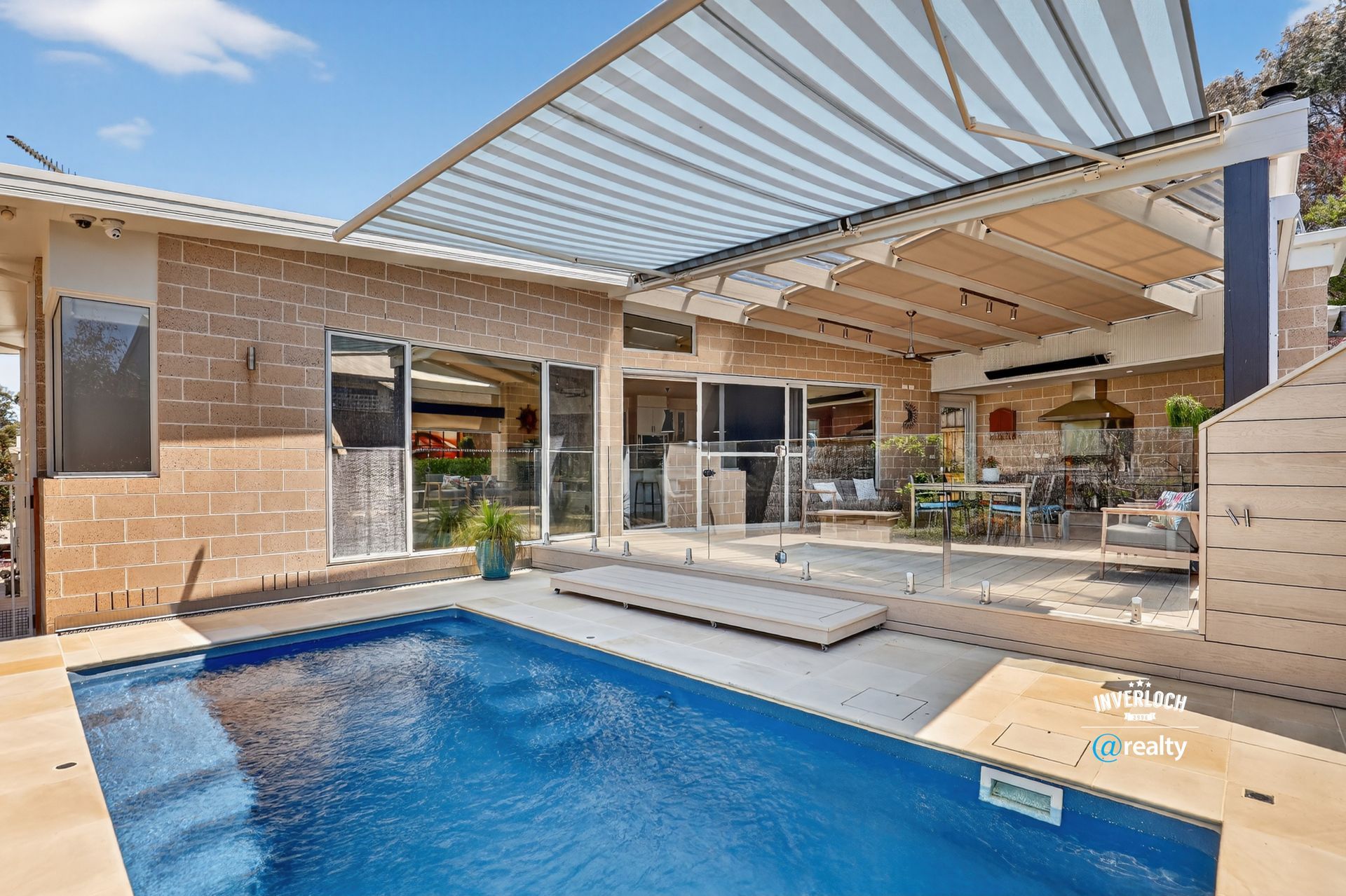 Poolside patio with retractable awning over a deck, brick house, and blue pool.