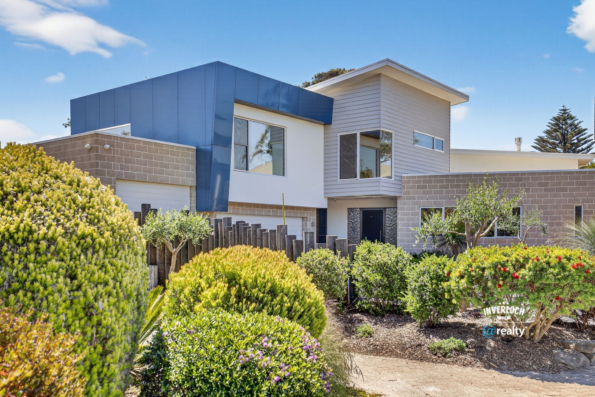 Modern two-story house with blue and gray exterior, surrounded by green shrubs and a small wooden fence.