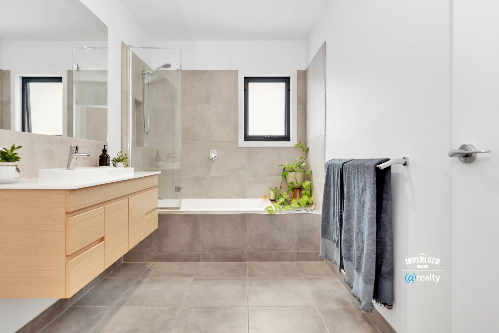 Modern bathroom with light wood vanity, gray tile, and glass shower. Gray towels hang on a towel rack.