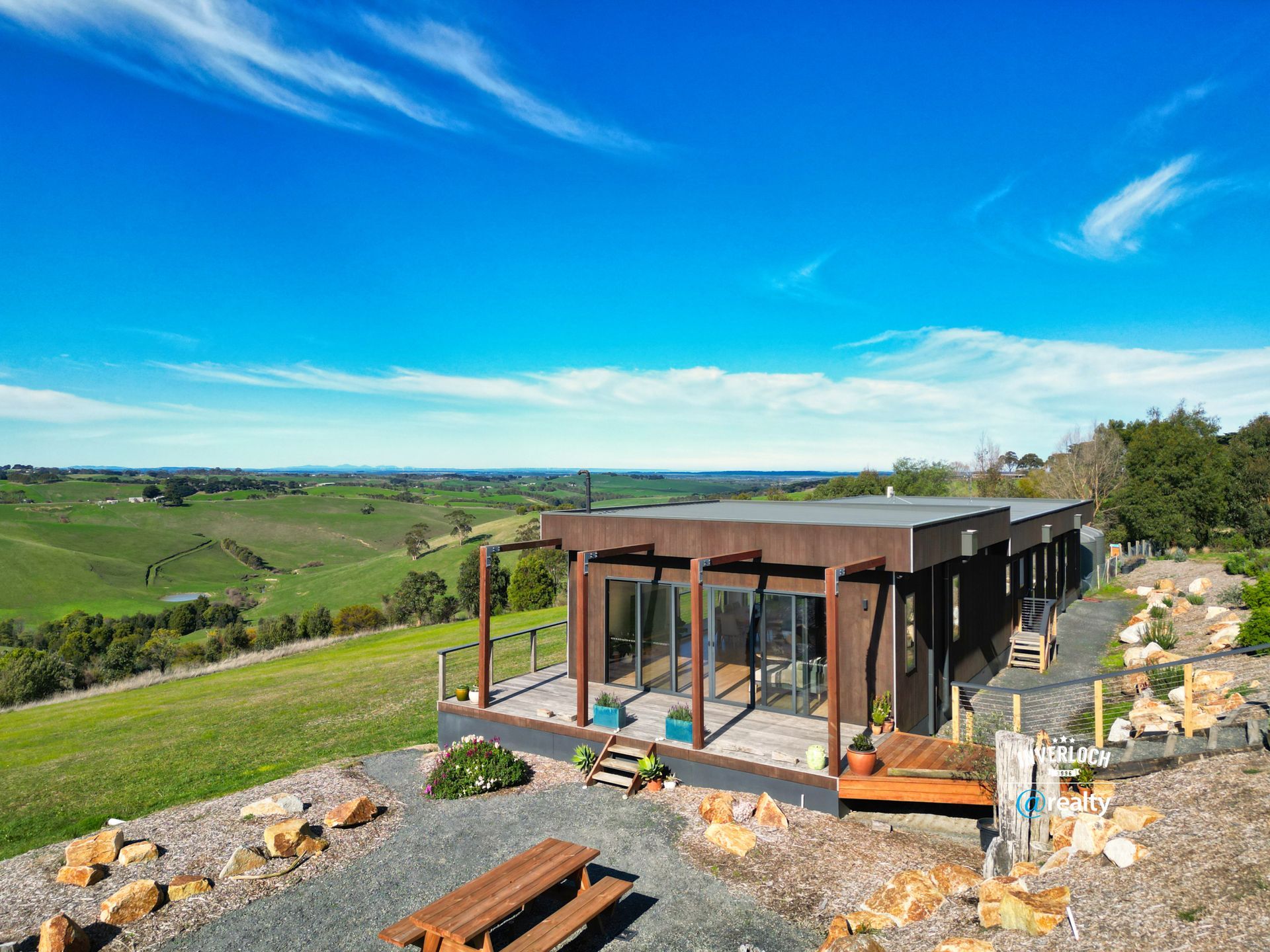 Modern brown house with a deck overlooking a green valley under a bright blue sky.