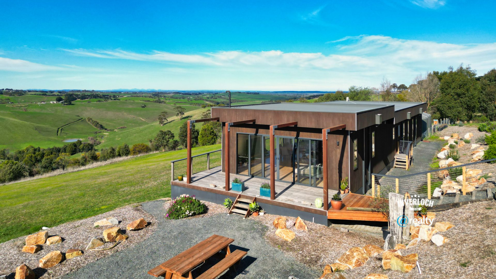 Modern glass cabin on a hillside with a deck, overlooking green fields and a blue sky