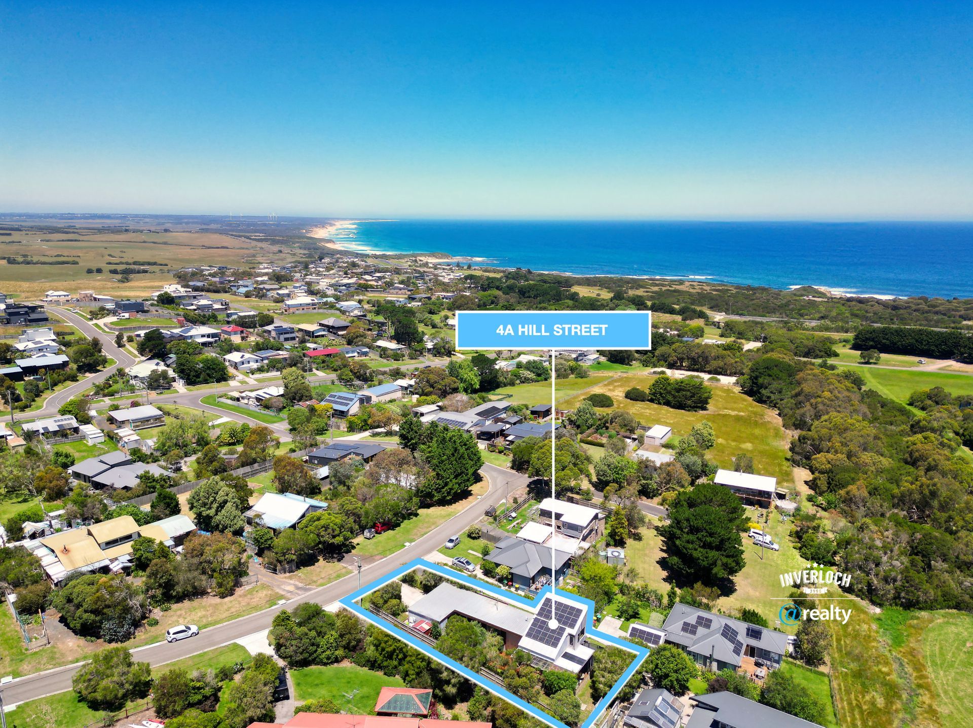 Aerial view of a coastal neighborhood with houses, trees, and a blue ocean horizon under a clear sky.