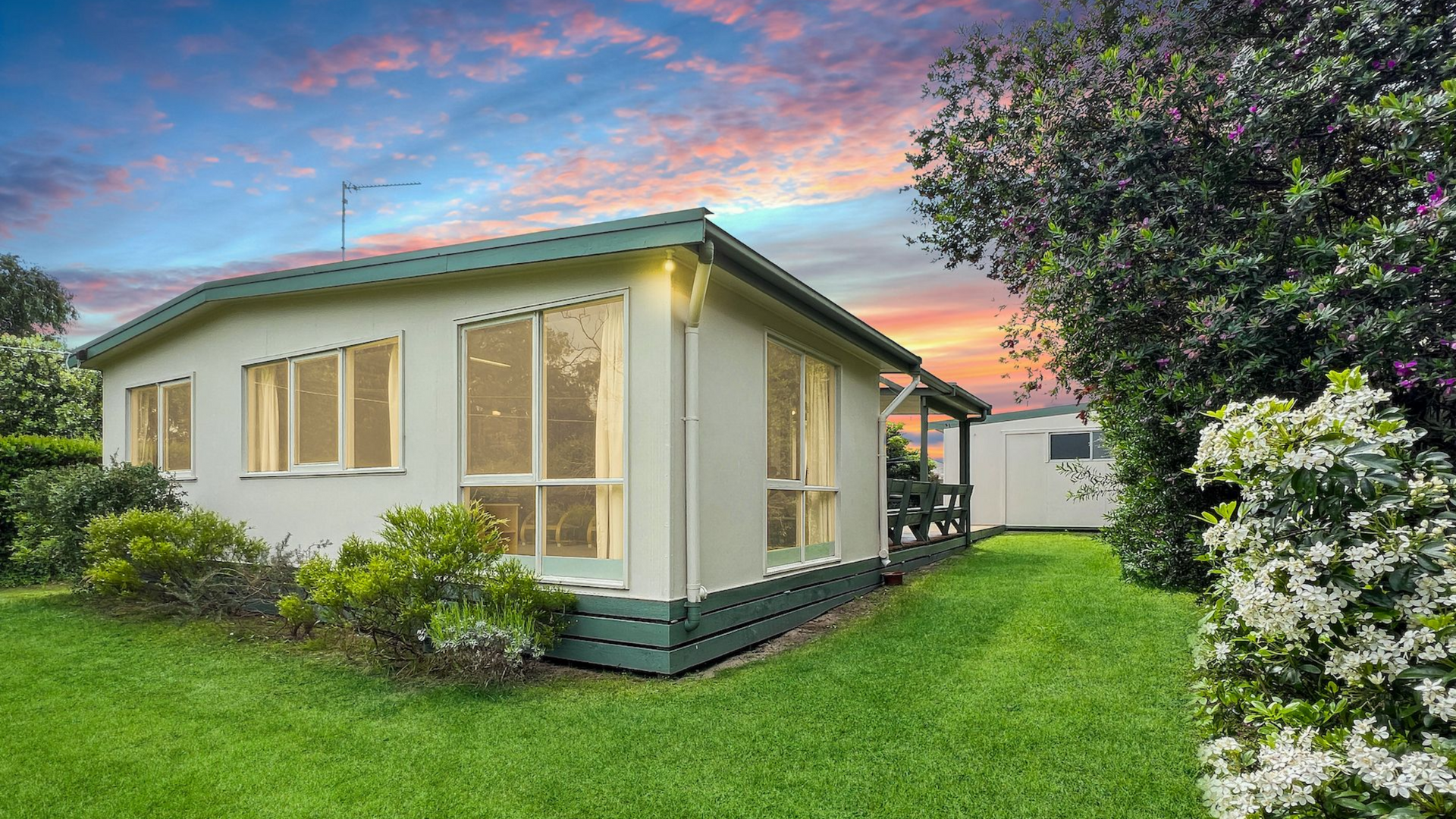 Small cream cottage with green trim in a lush garden at sunset, under a pink-blue sky