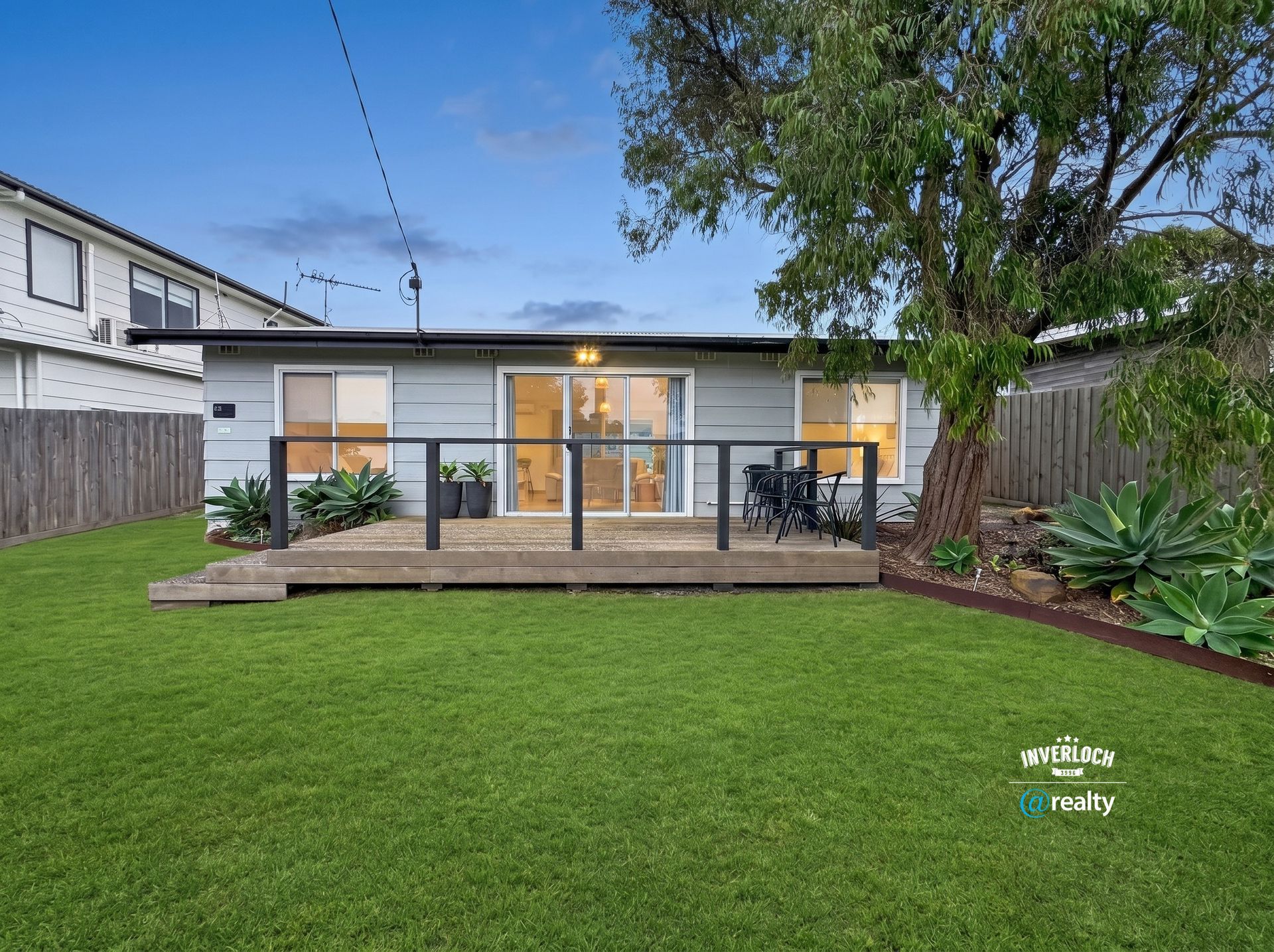 Backyard view of a modern single-story house with a wooden deck and large green lawn at dusk