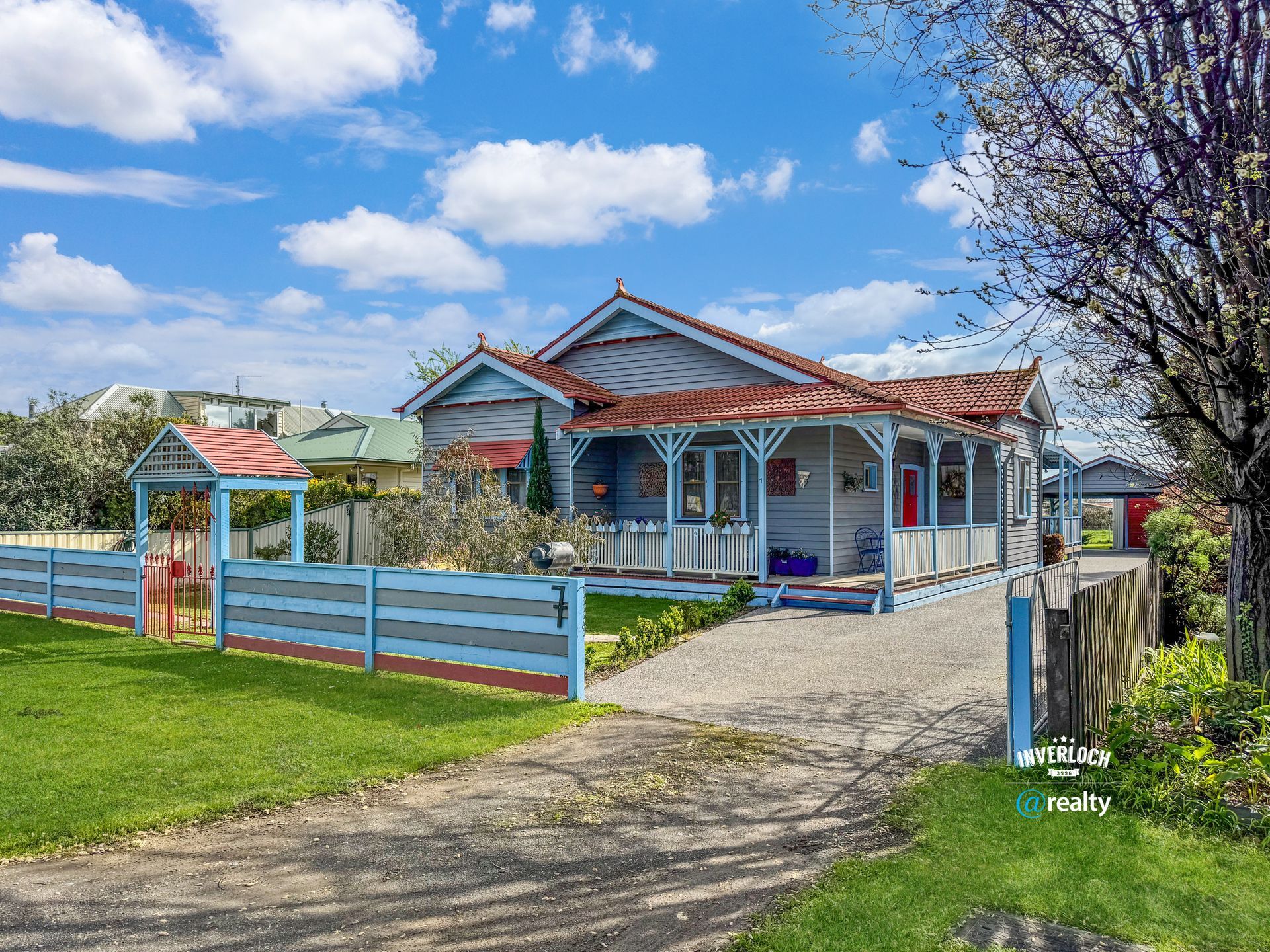 Blue house with red trim, white fence, and gravel driveway on a sunny day