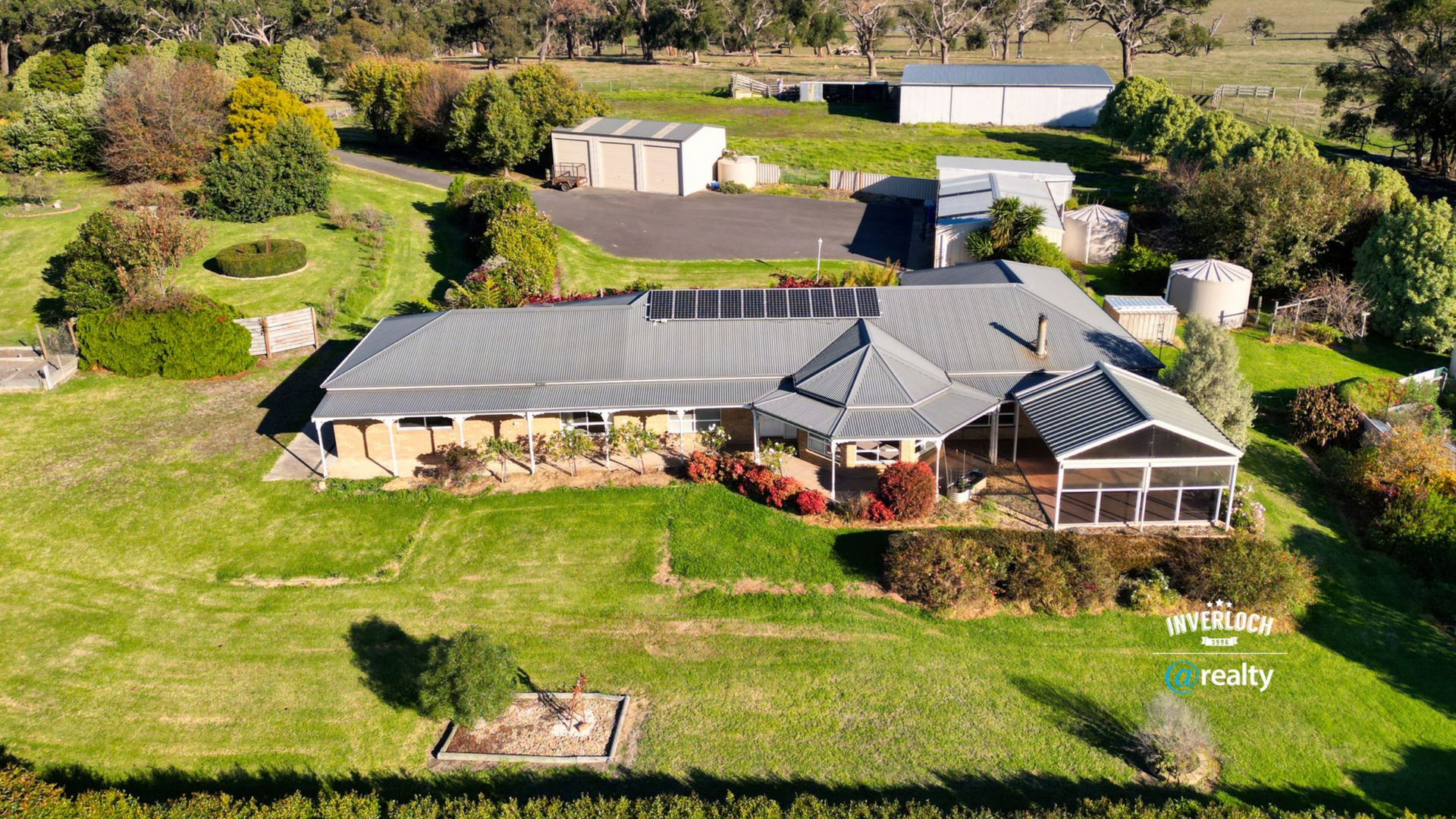 Aerial view of a countryside house with a dark roof, green lawn, trees, and outbuildings.