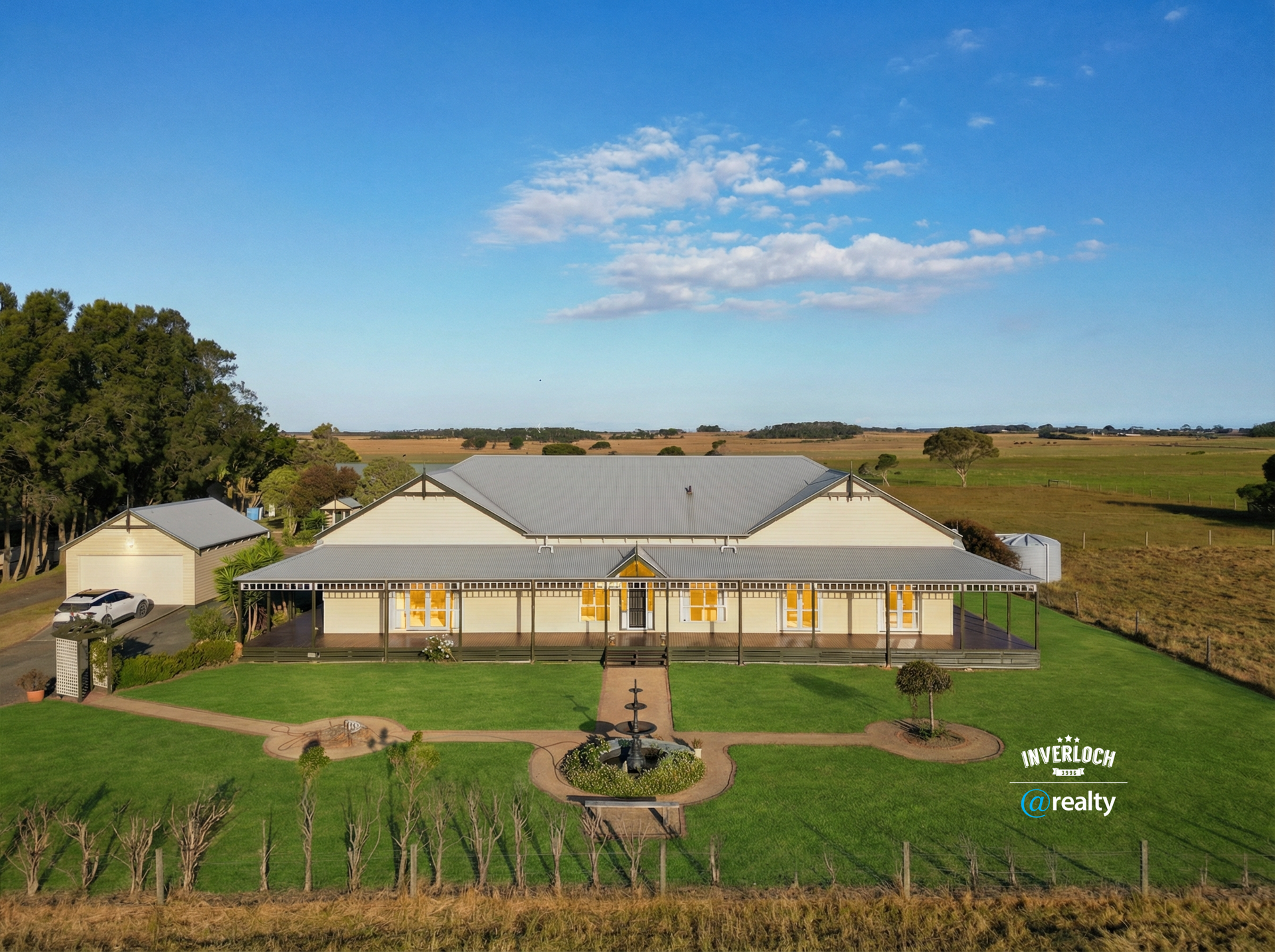 A large, single-story building with a wraparound porch and gray roof sits on a green lawn surrounded by farmland.