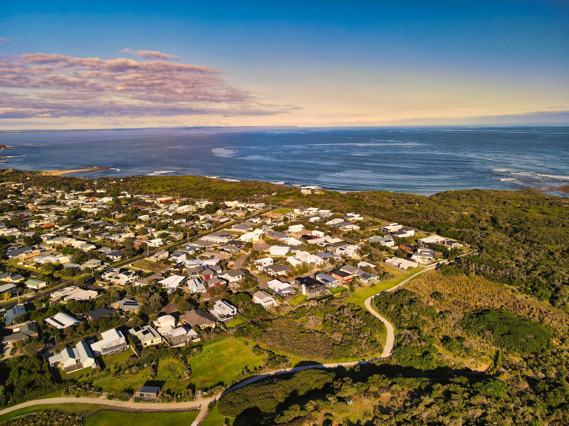 Aerial view of a Cape Paterson beside a blue ocean, with winding roads and green hills at sunset.