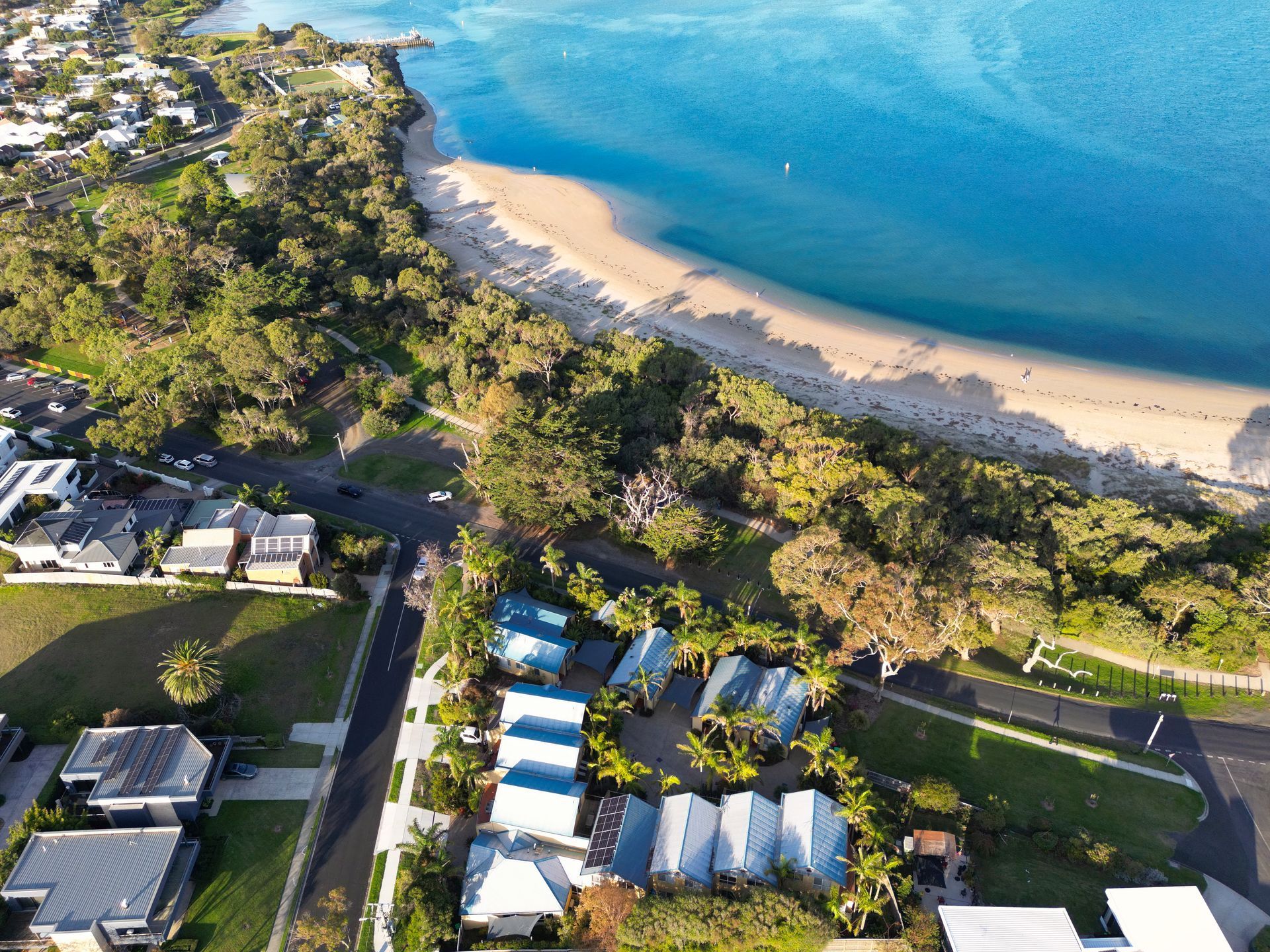 Aerial view of beach and ocean next to suburban homes and a row of blue-roofed buildings.