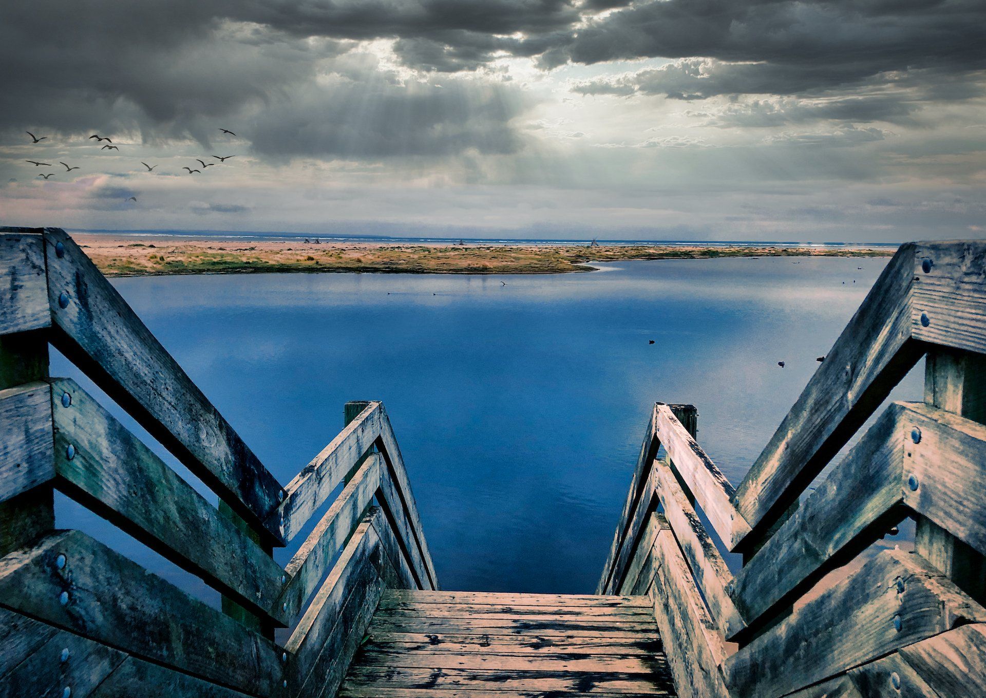 Wooden stairs descend to a calm blue lake under a dramatic cloudy sky