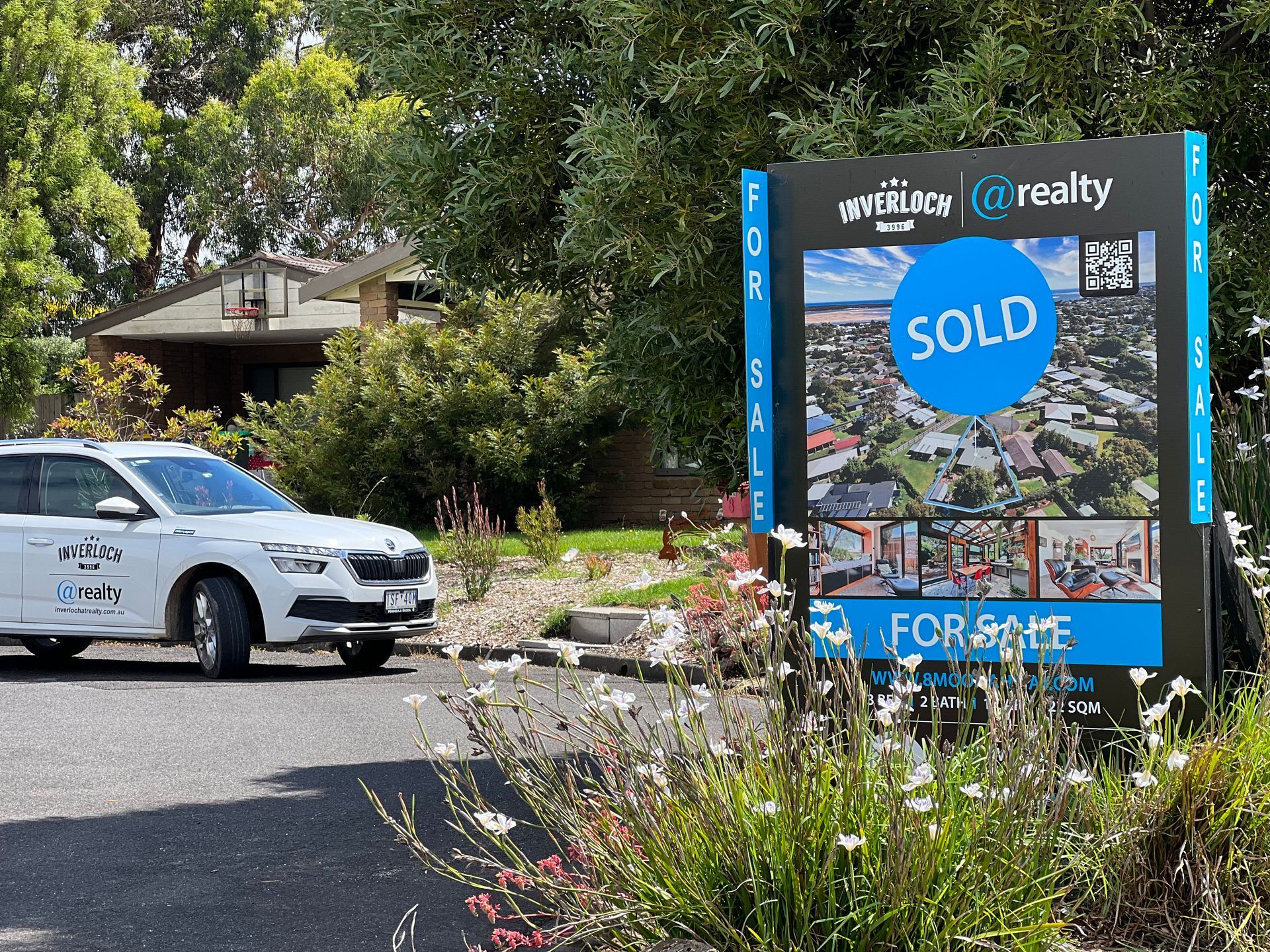 Real estate sign with blue “SOLD” and “FOR SALE” notices beside a white car on a driveway