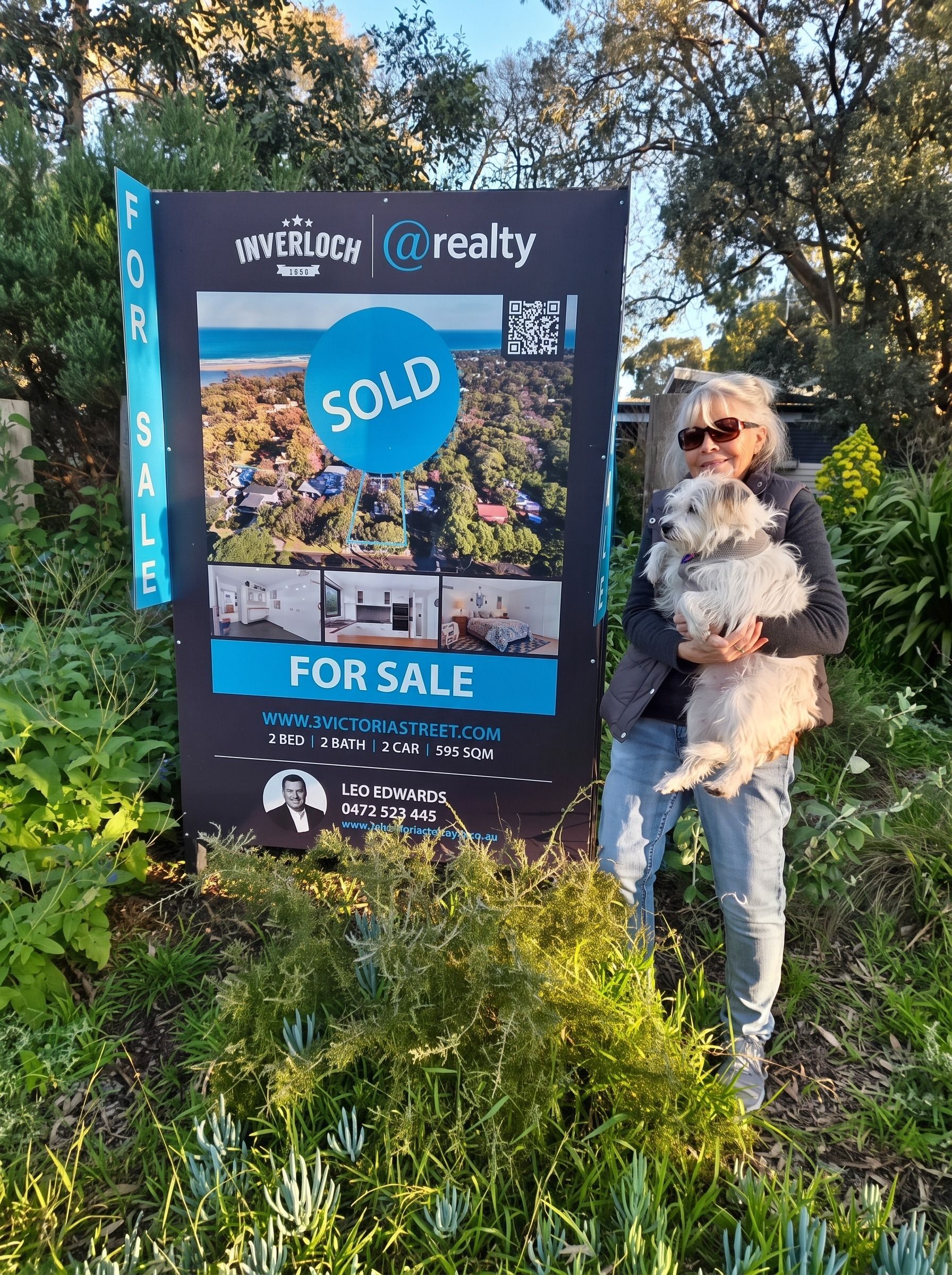 Person standing beside a real estate sign with a “SOLD” sticker and “FOR SALE” banner in a grassy area.