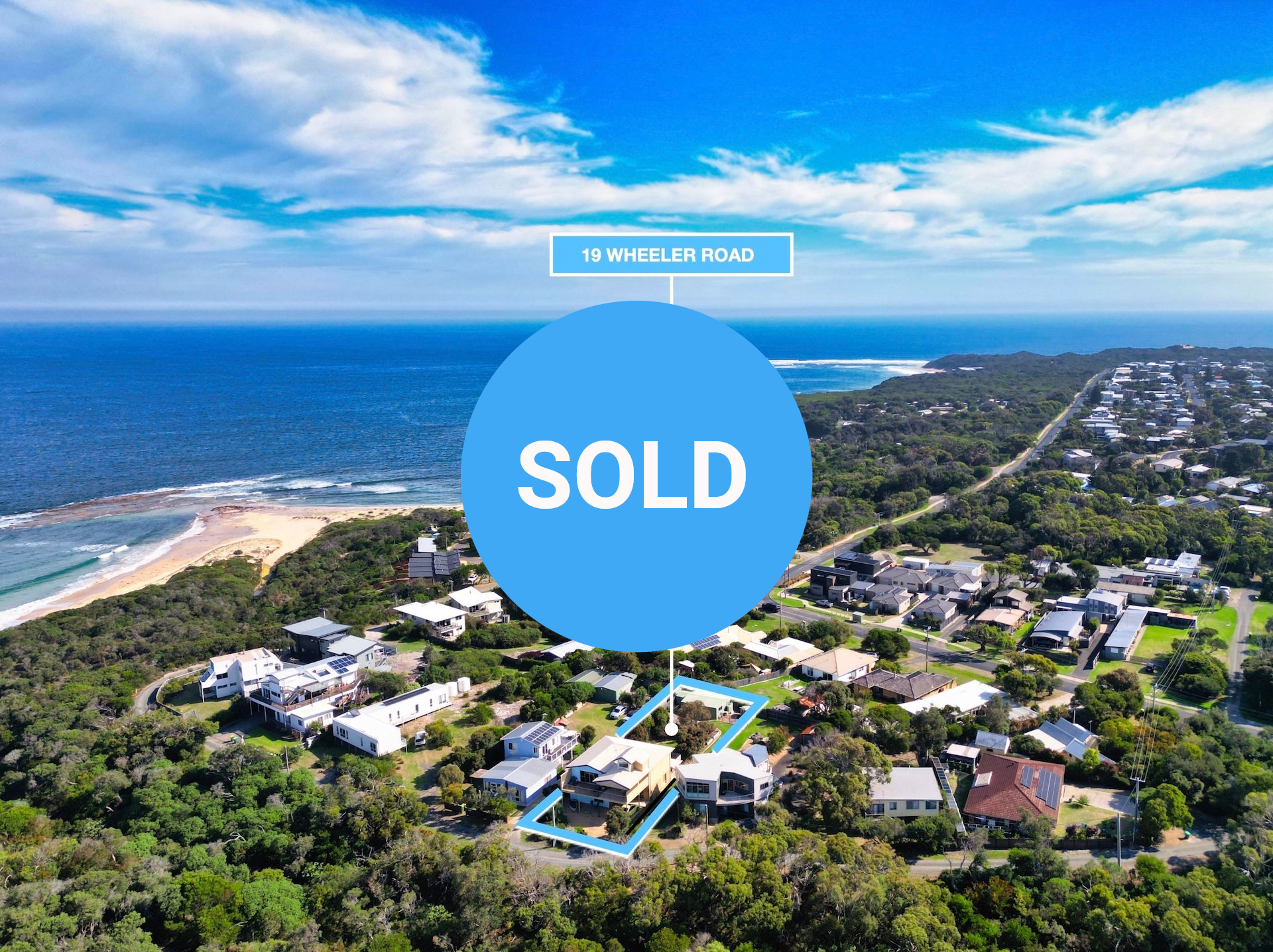An aerial view of a coastal neighborhood with a blue outlined property, located near a beach and ocean under a blue sky.