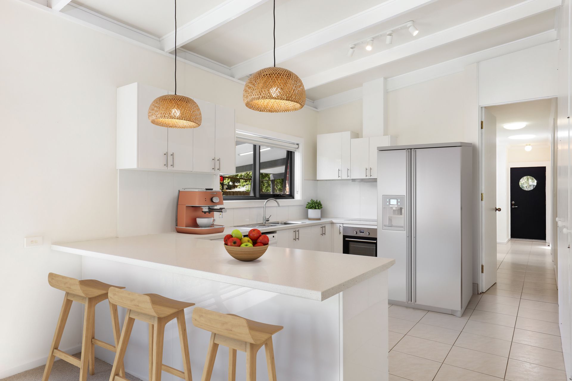 A kitchen with white cabinets , stools , a refrigerator and a bowl of fruit on the counter.
