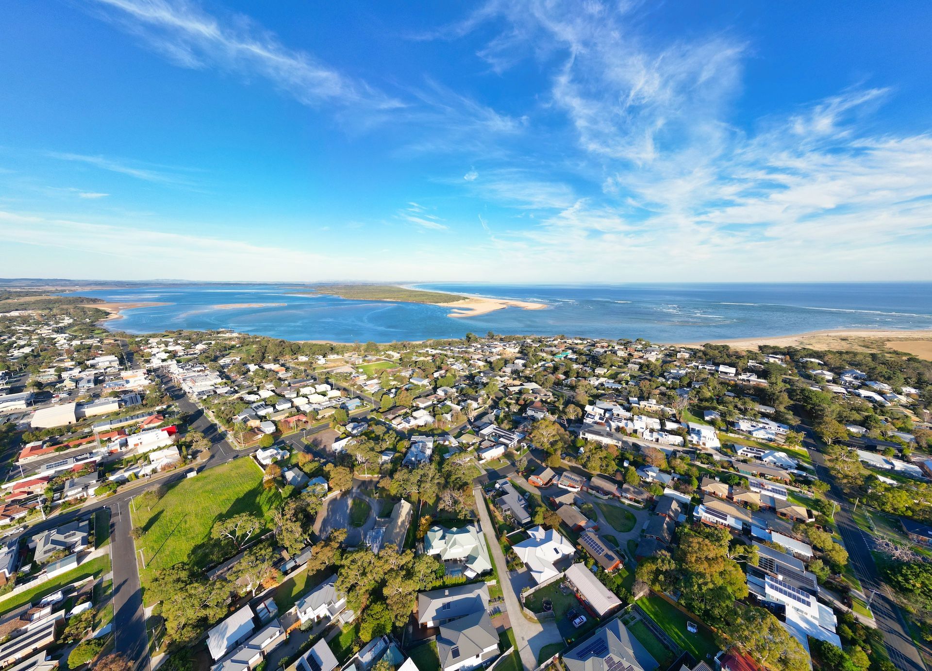 Aerial view of a coastal town with houses, green spaces, and a body of water under a blue sky.