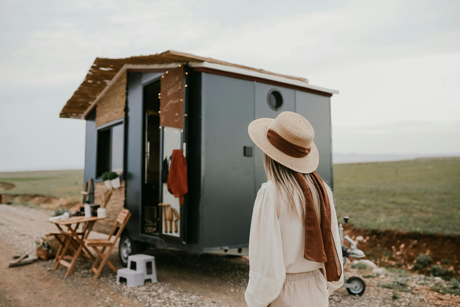 Woman in a straw hat looking at a tiny house on wheels, parked on a dirt road.
