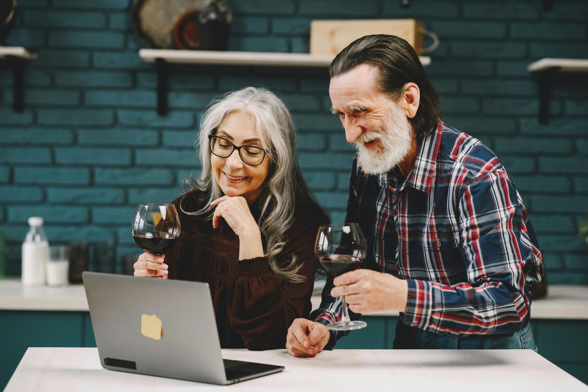 Older couple with wine glasses video chatting on a laptop in a kitchen.
