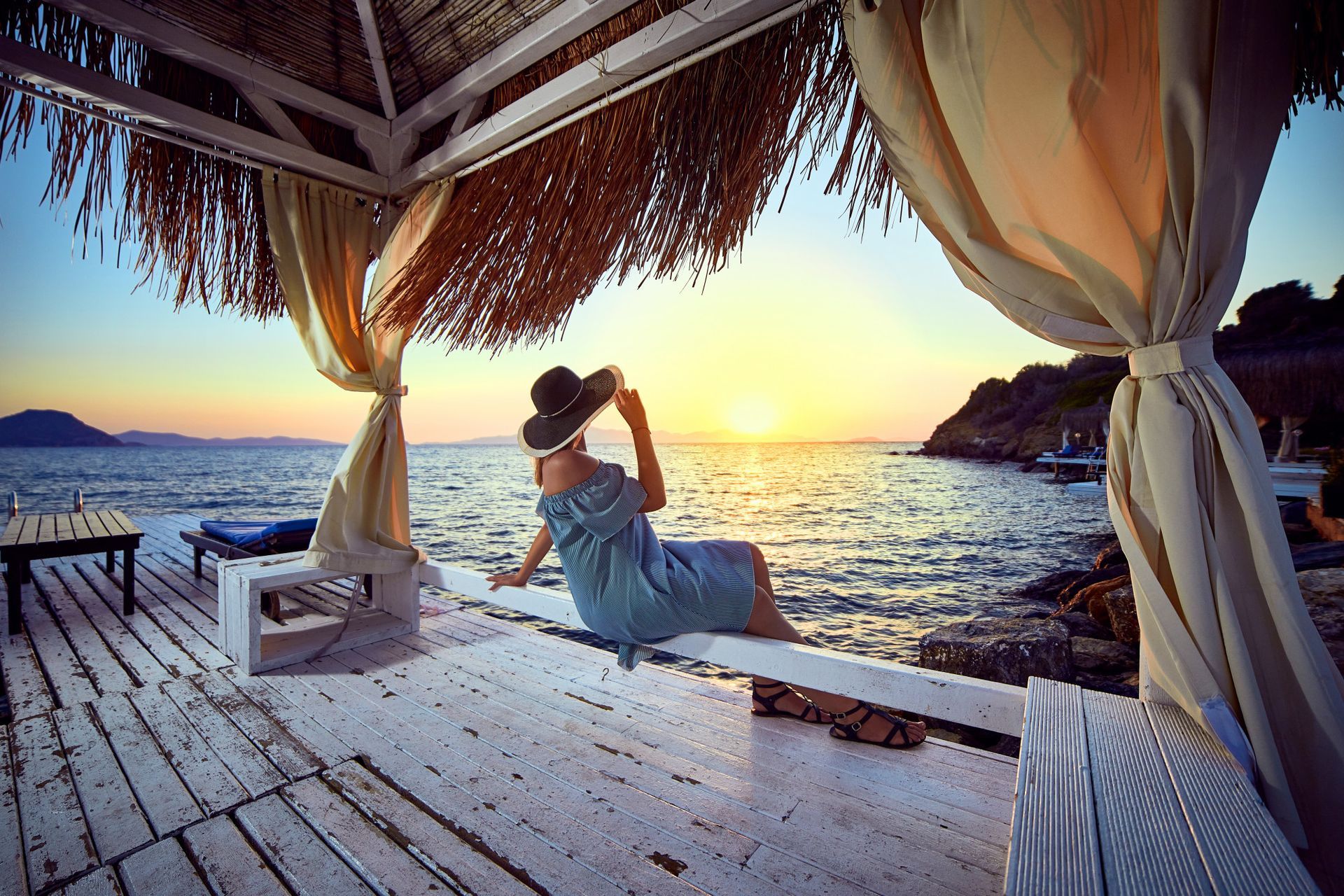 A woman is sitting on a bench under a gazebo overlooking the ocean at sunset.