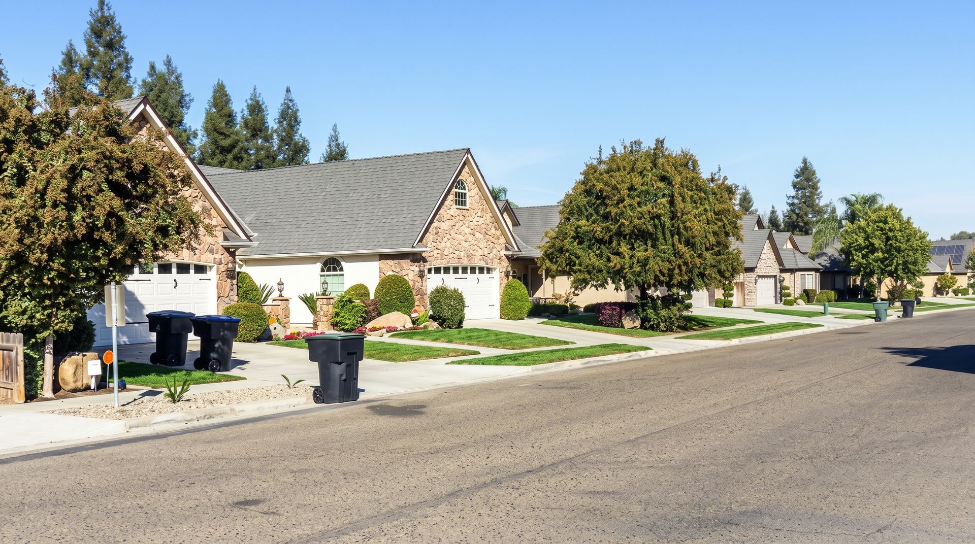 Suburban two-story beige house with dark roof, front porch, garage, and landscaped lawn on a sunny day