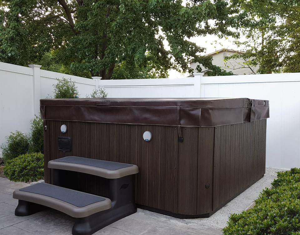 A dark brown hot tub with steps sits near a white fence, surrounded by greenery. Hot tub with brown exterior and steps, set next to a white fence and greenery.