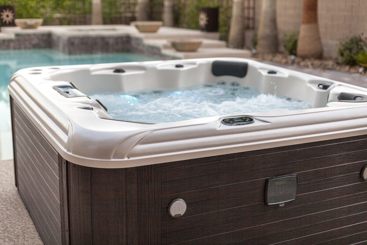 Hot tub outdoors with bubbly water, brown wicker-style siding, and a pool in the background. Hot tub with bubbling water outdoors near a pool.