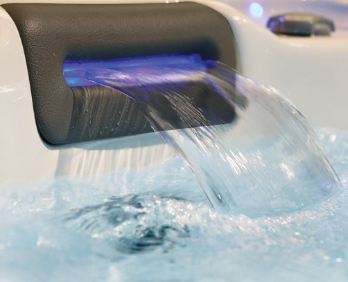 Close-up of a spa water feature with a blue light illuminating cascading water into a bubbly spa pool. Water cascading from a spout with blue LED lighting into a bubbly spa.