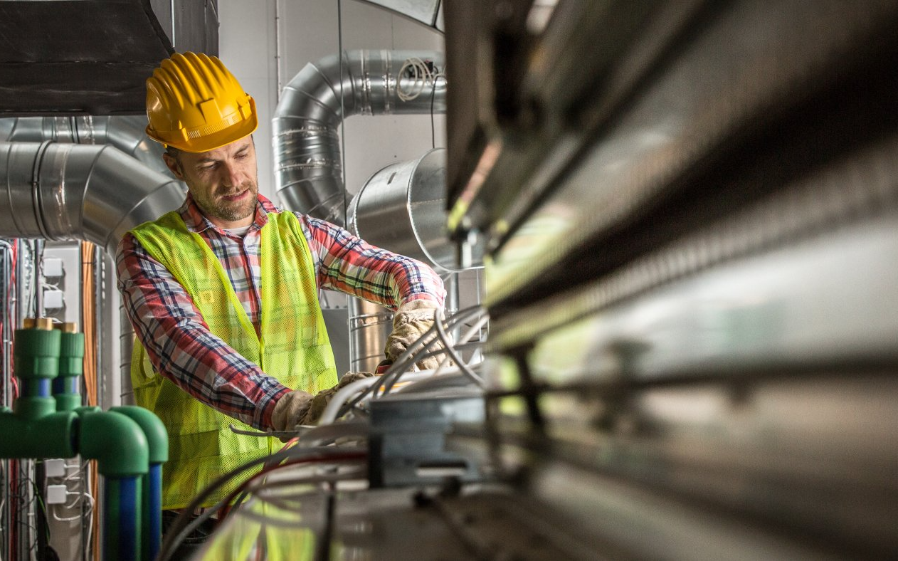 A man wearing a hard hat and safety vest is working on electrical wires in a factory.