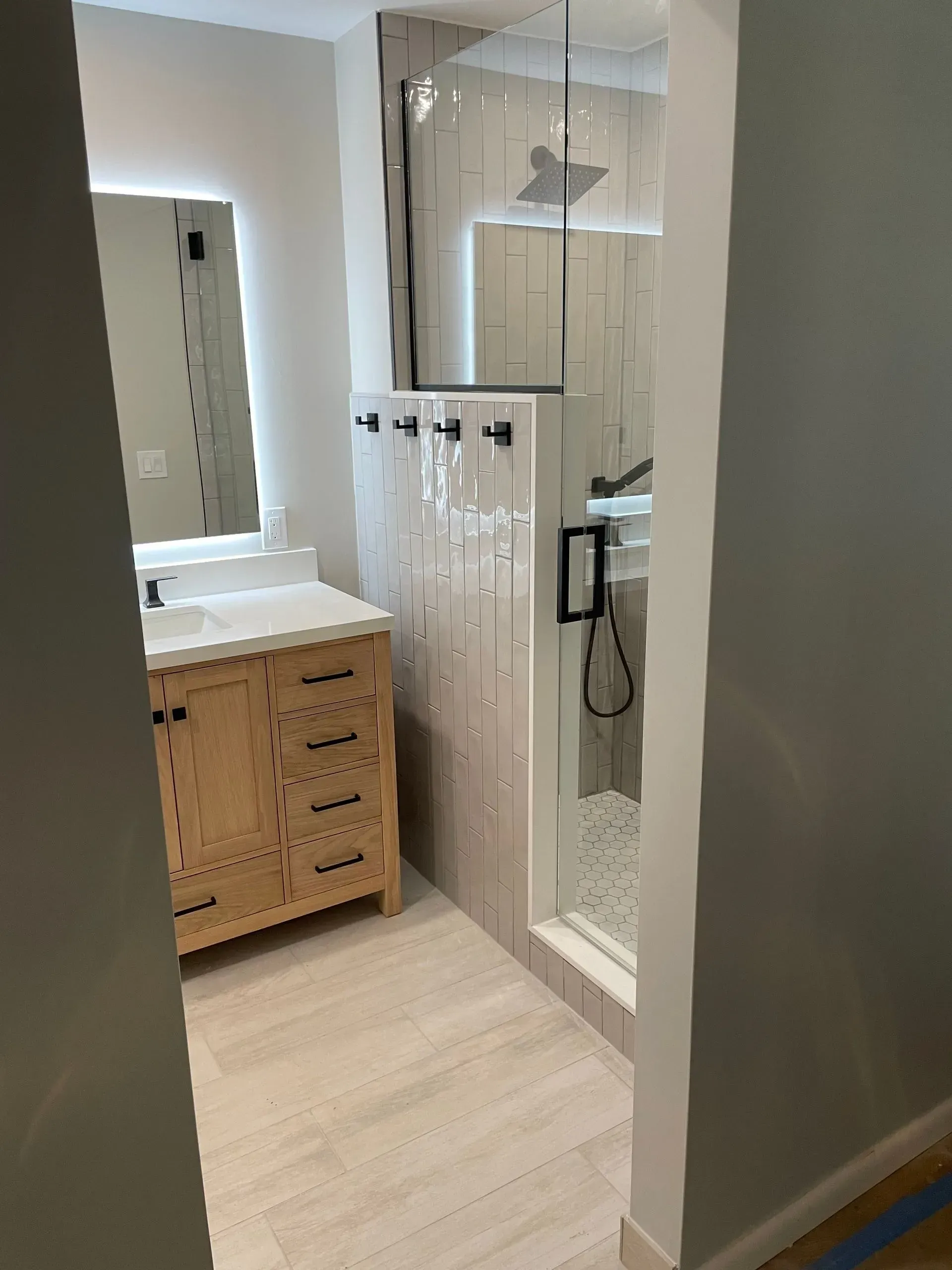 Small bathroom with wood vanity, mirror, glass shower, and light tile floor.