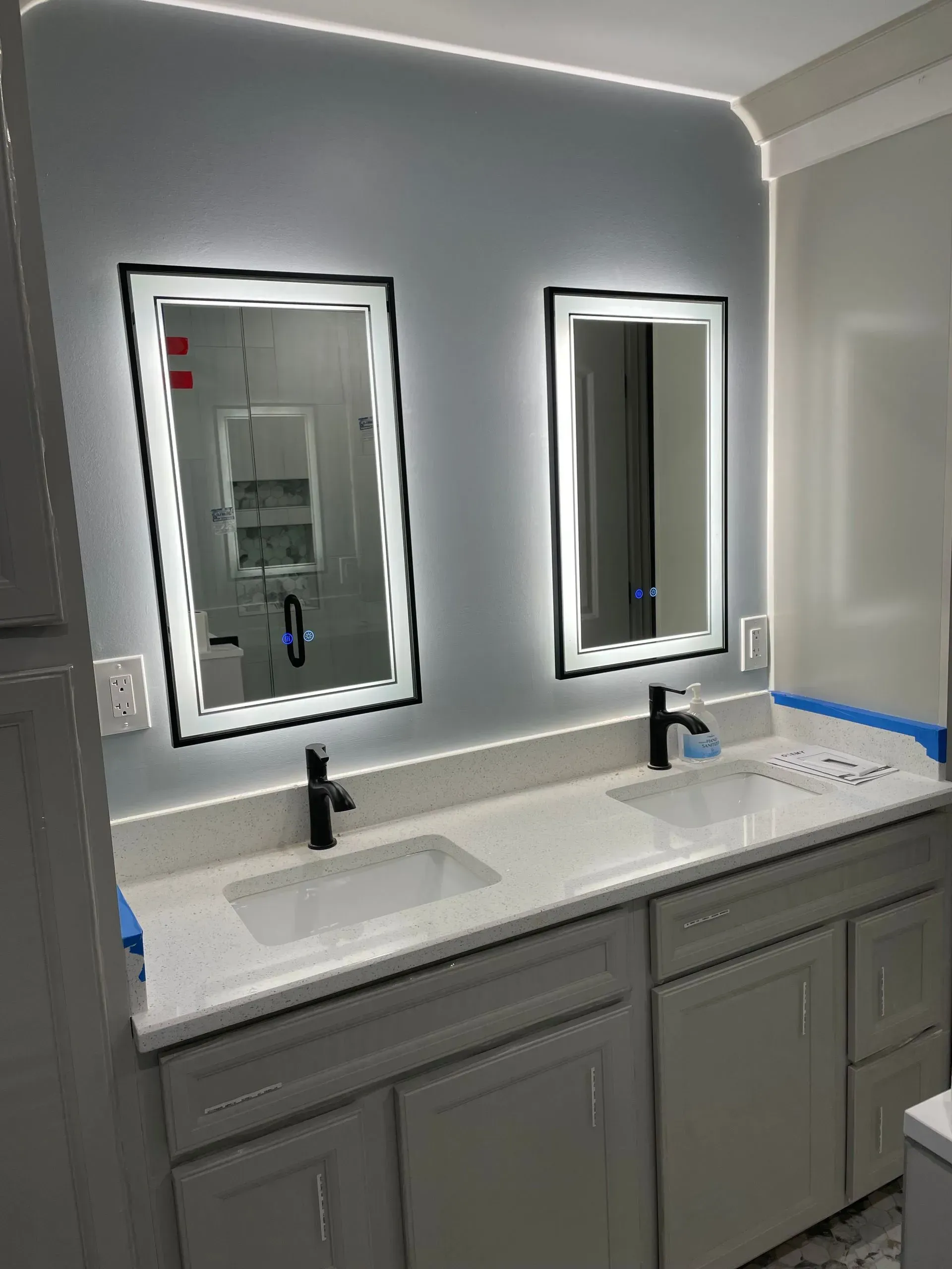 Modern bathroom vanity with double sinks, black faucets, and two illuminated mirrors above gray cabinets.