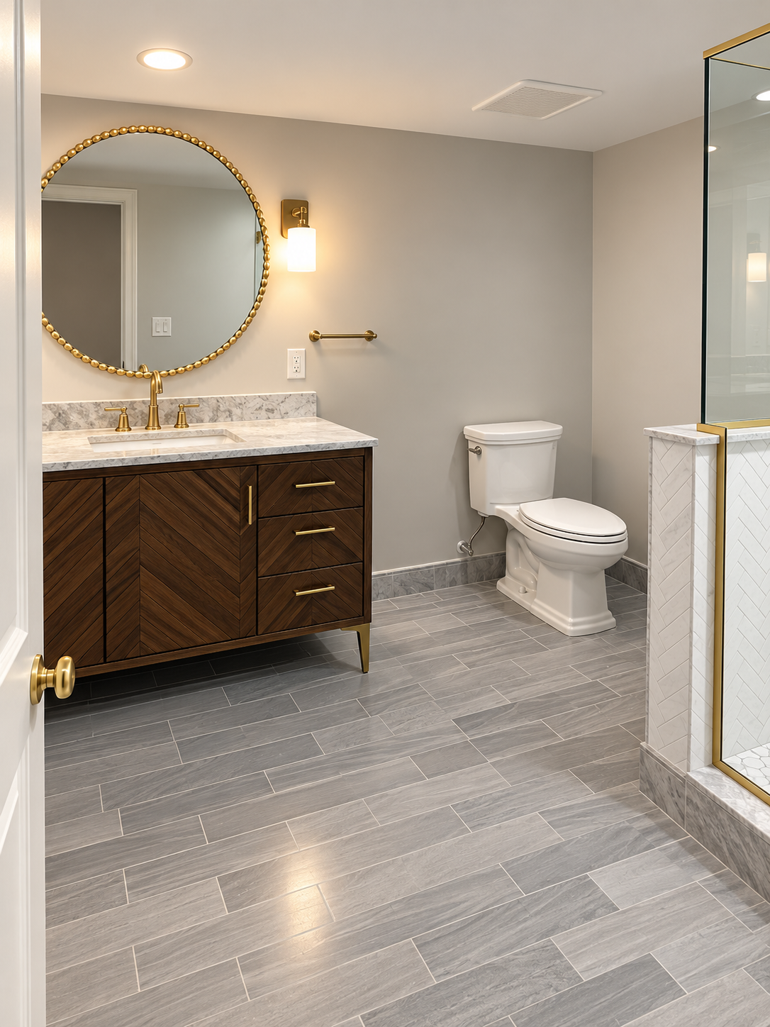Small bathroom with gray tile, round mirror above dark wood vanity, toilet, and glass shower door.