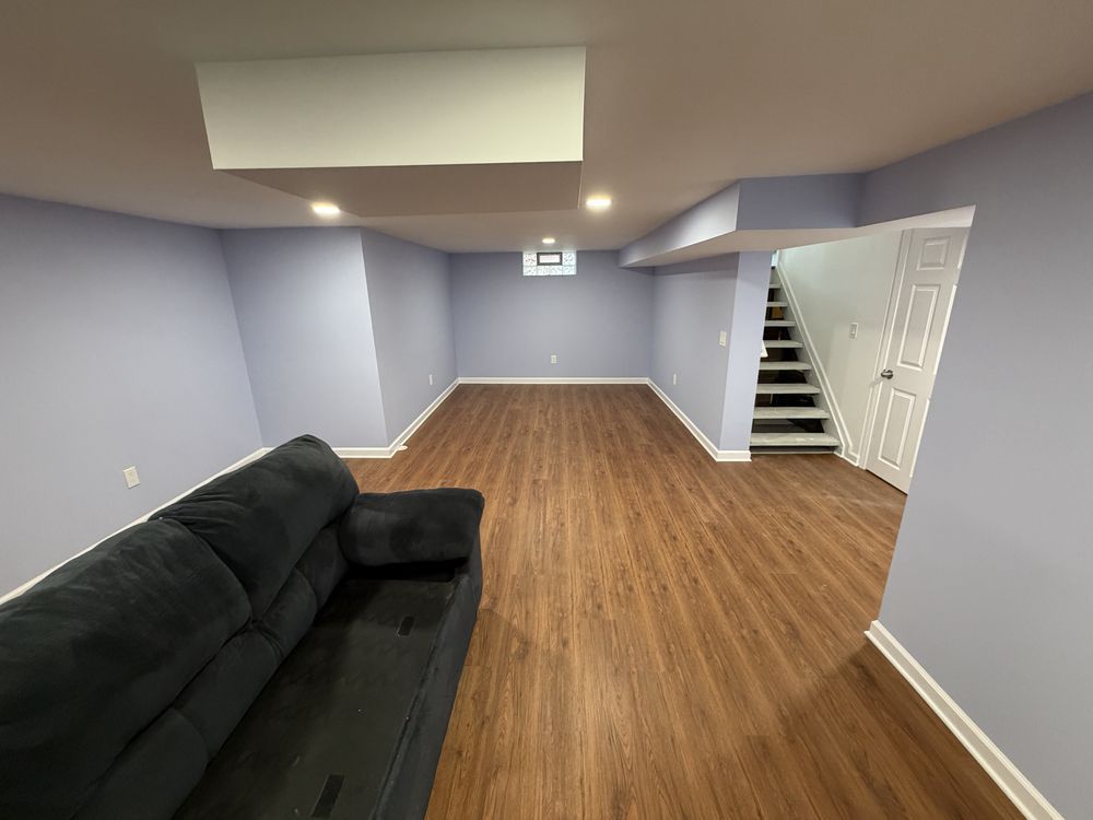 Empty basement living room with gray walls, wood-look floor, black sofa, and stairs by a white door