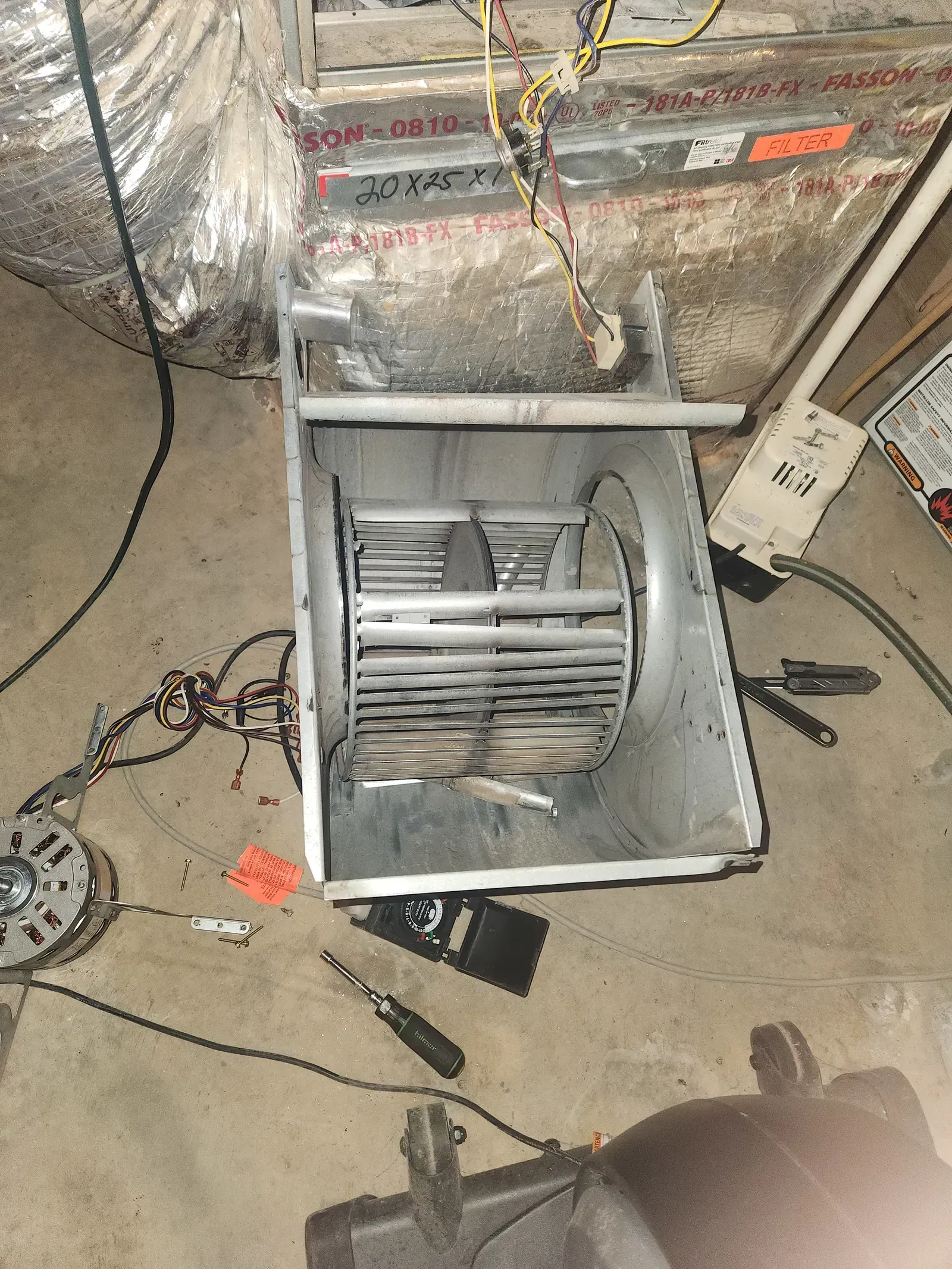 Furnace blower assembly, disassembled. Metal fan blades inside a housing, tools on the concrete floor.