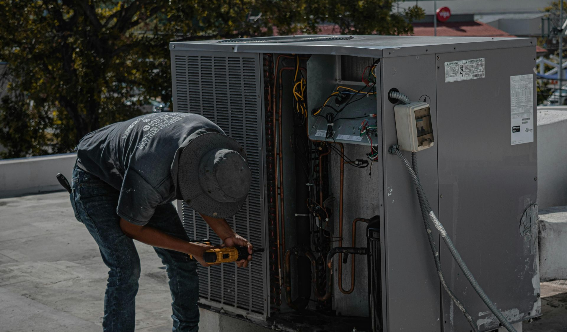 HVAC technician repairs outdoor air conditioner on a rooftop. Man in gray clothes uses a screwdriver.