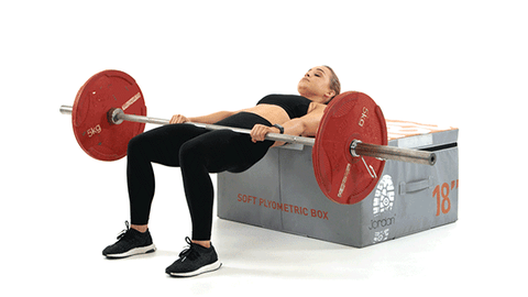 Woman doing barbell hip thrusts on a box. She has black leggings, a sports bra, and sneakers.
