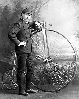 Man in dark suit and cap poses with a high-wheel bicycle.
