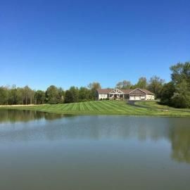 A lakeside house with a green lawn reflected in calm water under a clear blue sky.