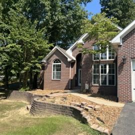 Brick house exterior with arched windows, a front walkway, and a low retaining wall in a wooded yard
