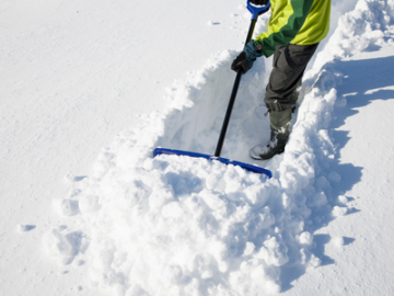 Person shoveling snow with a blue shovel on a snowy surface, wearing a yellow jacket.