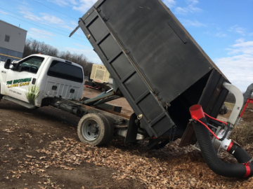 Tow truck lifting a black dumpster trailer, with a white truck nearby on a leaf-covered lot.