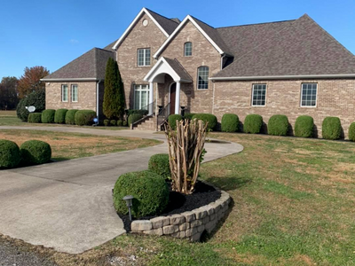 Large suburban brick house with a curved driveway, landscaped lawn, and trimmed bushes
