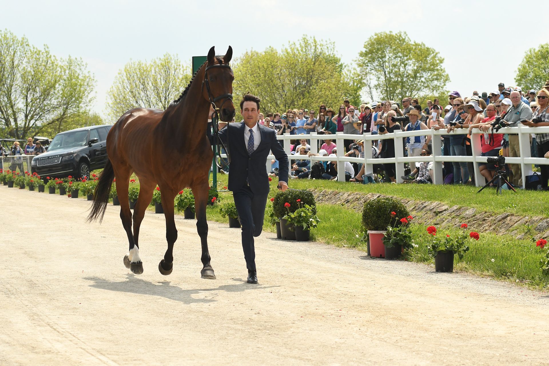 A man in a suit is leading a brown horse on a dirt road.
