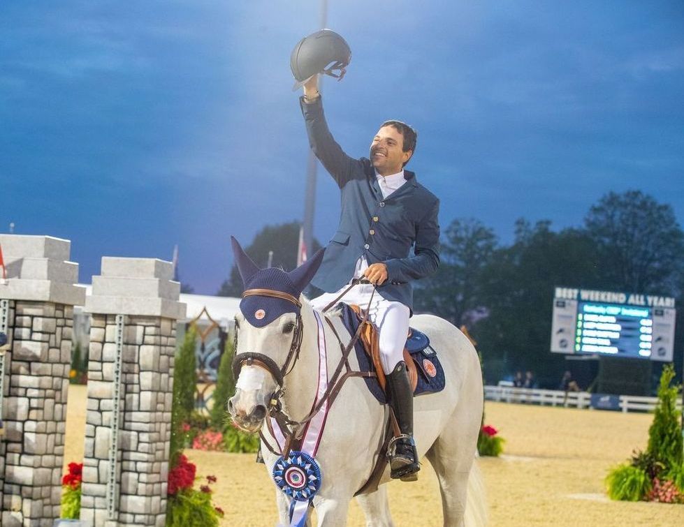 A man is riding a white horse and holding his helmet in the air